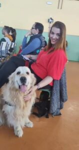 Foto de Isabel entre el público, sentada en una silla junto a Luca, su Perro de Asistencia para Discapacidad Física.